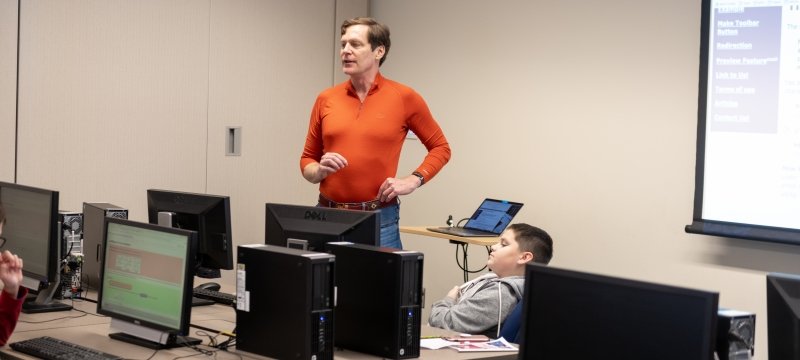 A man teaching in front of a computer class in a bright orange shirt with a young boy facing him and computer monitors with a white screen in the background