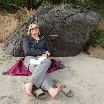 A woman sits in the sand while leaning against a large boulder. An open book is in her lap. She is smiling.