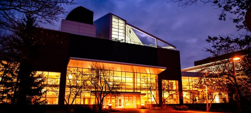 The Dow building on the Michigan Tech campus glows with light just before dawn. The sky is somewhat cloudy in the background and the building is surrounded by trees.