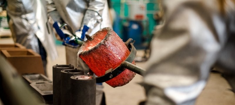 Two people in fire-resistant clothing pour melted metal from a red-hot crucible. 