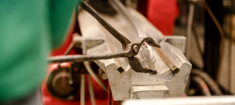A person uses a pair of metal tongs to pull an aluminum rod through an extruder machine.