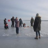 A group of people of all ages stand around a hole drilled into the ice.
