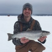 A man kneels on the ice holding a silvery, polka-dotted fish.