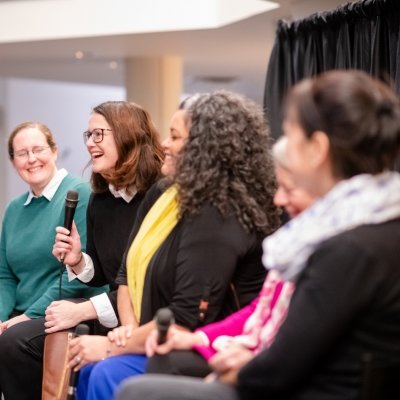 Four woman sitting and laughing in a theater lobby during a talk. One woman is speaking into a microphone. The woman sitting next to her is looking at her, holding a microphone in her lap, a woman in profile is listening and smiling and the other two women are in soft focus.