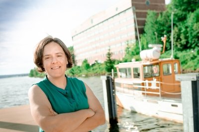 A woman stands on a dock smiling on a sunny summer day with a blue sky, the Keweenaw waterway, and a research vessel in the background in northern michigan.