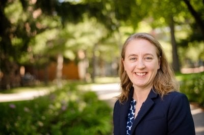 A woman wearing a blazer smiles with green campus trees and brick buildings in the background on a sunny day outside.