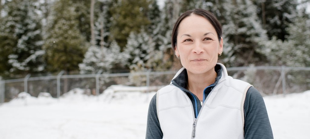 A woman smiles in the snow wearing a white vest with a fence, drifts, and snow-capped evergreen treeline behind her.