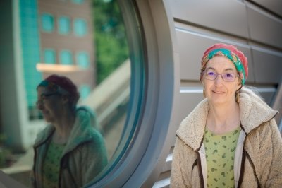A woman wearing glasses and a colorful cap is reflected in a porthole window of a building outside in winter at a University's waterfront research center.