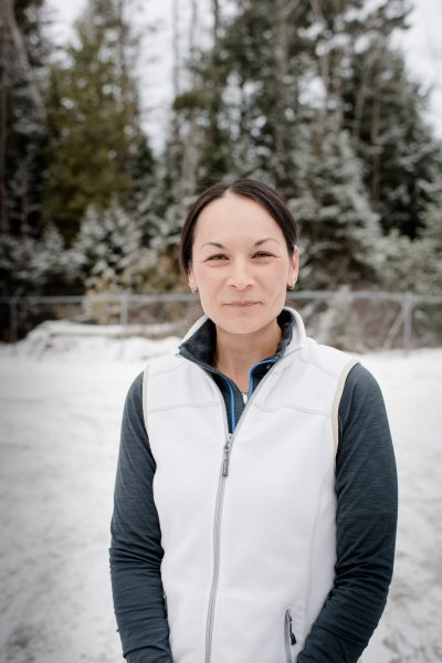 A woman smiles at the camera with snowdrifts, a fence, and snow-tipped evergreens in the background on a winter day with blue sky.