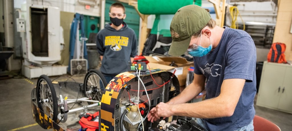 Students wearing face masks and safety goggles work on an engine in a garage space, one with his hands on the engine and the other wearing a Michigan Tech sweatshirt looking on next to the wheels. There is a roll of green gauze behind them in a shop with a fan and industrial equipment in the background.