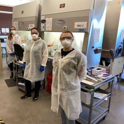 Three people who are wearing masks, protective eyewear, and a lab coats stand in front of the vent hoods in the COVID-19 lab at Michigan Tech.