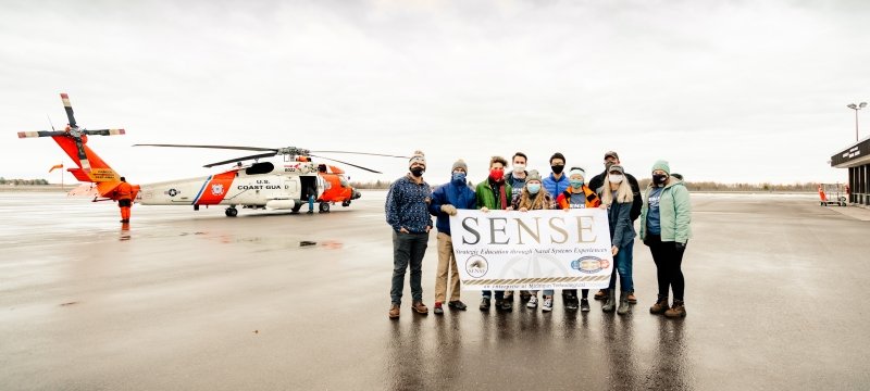 Eight students stand outside at an airport on wet pavement carrying a SENSE Enterprise banner with a US Coast Guard Helicopter behind them on a fall day with a gray sky.