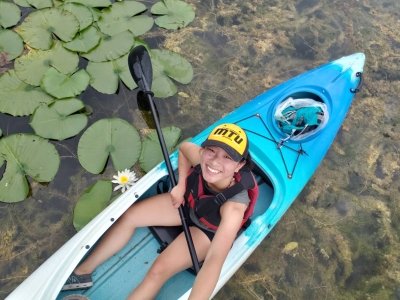 A young woman wearing an MTU cap smiles up from a kayak with lilly pads and one lily on the water as she holds her paddle and is wearing a personal flotation device.