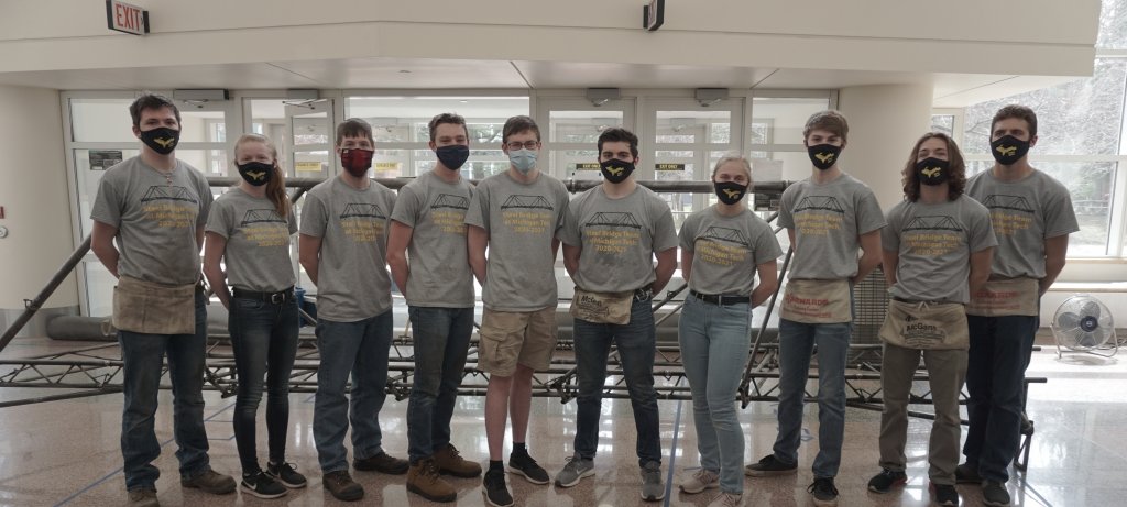 10 young people wearing Michigan Tech Steel Bridge Team t-shirts stand in front of the bridge they assembled indoors with glass doors and windows behind them on a college campus. They are wearing face masks and some have tool belts.