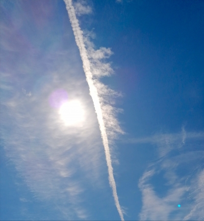 Blue sky with a long white contrail curving through a sun flare.