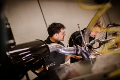 Two people with safety glasses on stand in a room full of metal equipment and plastic hoses.