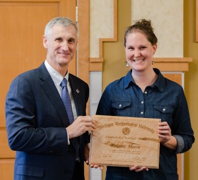 A smiling woman stands with a smiling man in a suit. They are holding a wood plaque that says Brigitte Morin Distinguished Teaching Award