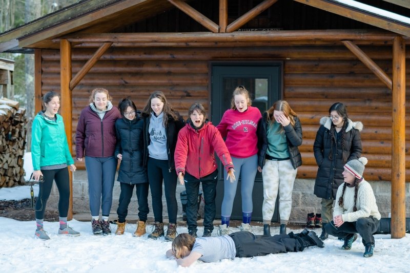 A line of students clowns around in front of a log cabin in the snow during a nature immersion in Upper Michigan.
