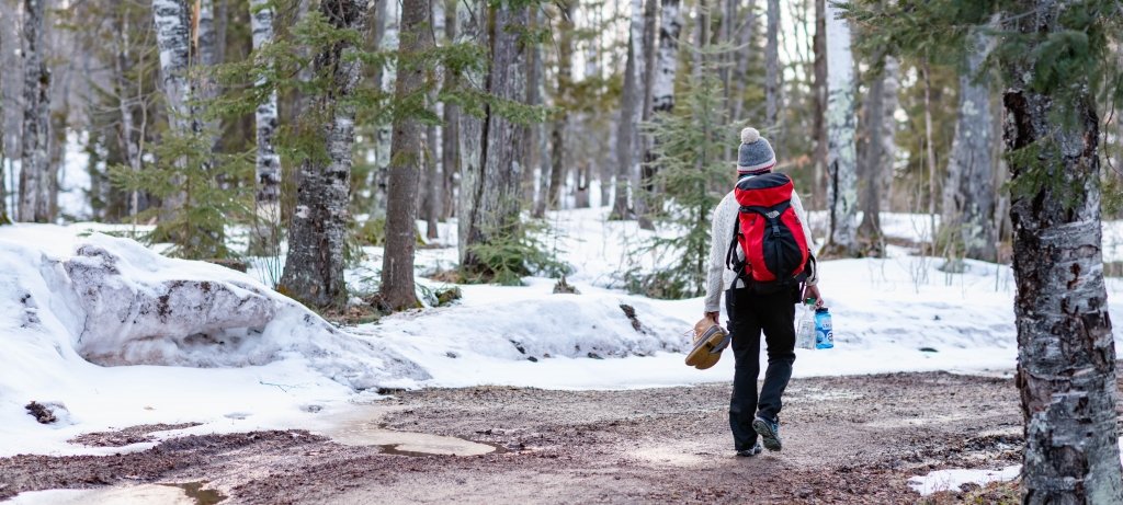 A young woman with a backpack and her back to us carries her snow boots and water on a trail back to a nature psychology exploration in a wilderness park.