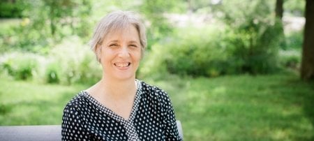 A woman smiles with a green outdoors background with a college campus in soft focus behind her. She is a researcher and faculty member.