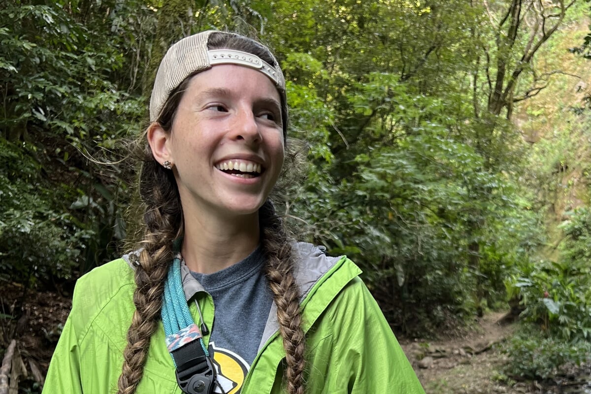A young environmental engineer with braids and a Michigan Tech Husky shirt smiles in Costa Rica near a running stream.