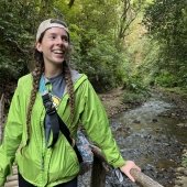 A young environmental engineer with braids and a Michigan Tech Husky shirt smiles in Costa Rica near a running stream.