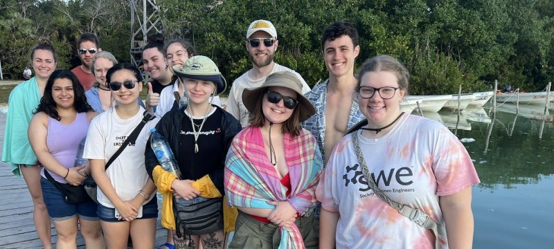 Twelve young people from Michigan Tech smile on a dock with water and small boats behind them as they study abroad in the Yucatan Peninsula.