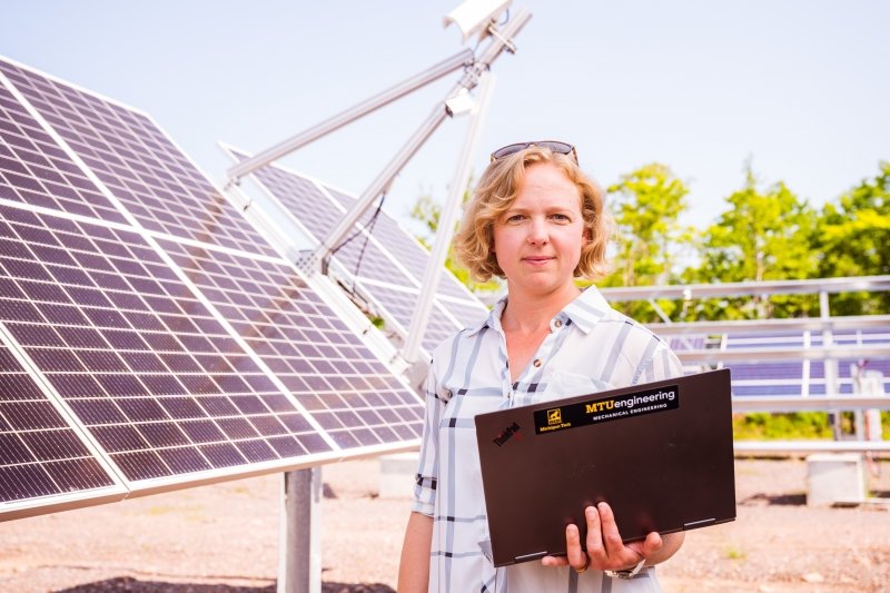 A researcher holding her laptop that reads Michigan Tech Engineering stands in front of a solar panel array as she conducts research as the lead of Michigan Tech's Great Lakes Energy Group