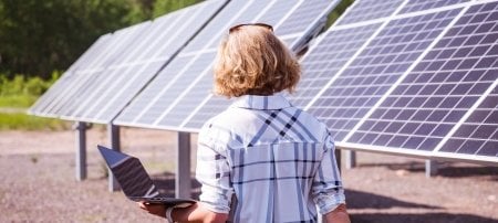 A woman with her back to the camera walks along an array of solar panels, holding a laptop computer she is using to observe data.