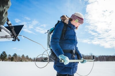 Cora Mullins wears a backpack spectrometer and extends the measuring apparatus with both hands over the snow on a clear day.