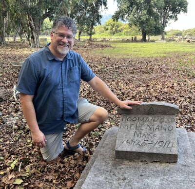 Joel Neves kneeling next to a gravestone with "Kia Aoomanaono Mele Mano Make Jan 25 1914" carved on it.