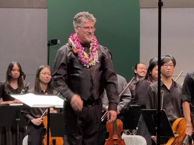 Joel Neves wearing several leis in front of a section of the orchestra.
