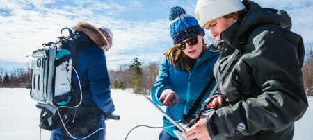 Three Michigan Tech researchers collect data on a snowy field site using a laptop computer and backpack spectrometer.