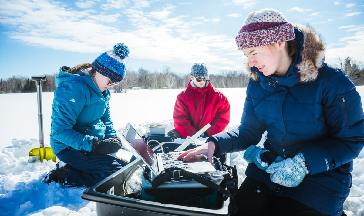 Three researchers work from a snowy field site with a sled full of equipment and instruments, including laptop, shovel and portable spectrometer.