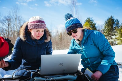 Researchers wearing winter coats, hats and snowpants work from a laptop computer in a field of snow.