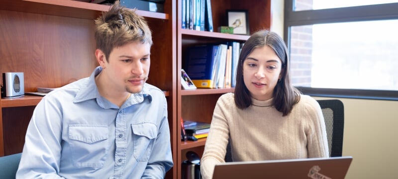 From left, Michigan Technological University research duo Nathan Price and Anna Stuhlmacher are seated side by side at a table, looking at a laptop.