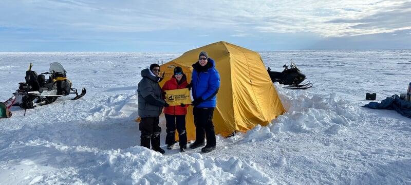 Researchers holding a small Michigan Tech flag standing outside a tent on the ice with a couple snowmobiles.