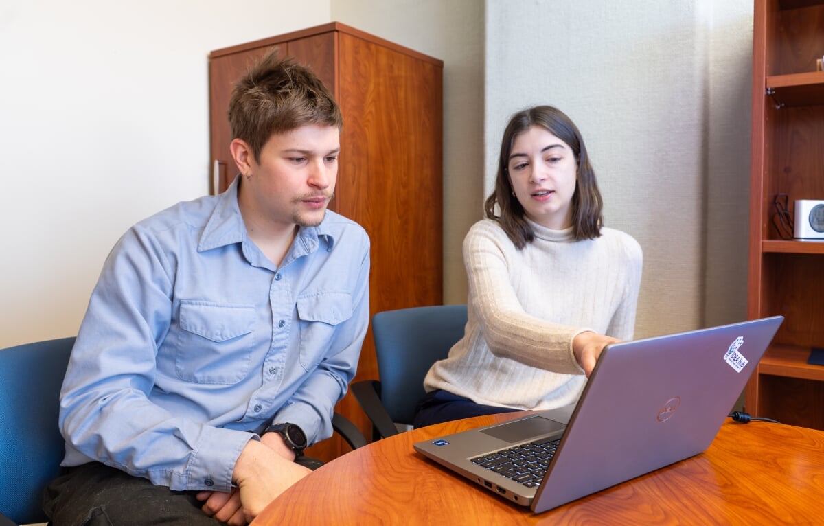 Left to right, Nathan Price and Anna Stuhlmacher are seated side by side at a table, looking at a laptop computer screen.
