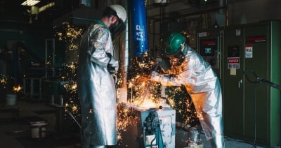 Two foundry workers, clad head-to-toe in heat-protective gear, work on a metal-casting project in MTU's foundry, with flying sparks and glowing hot metal.
