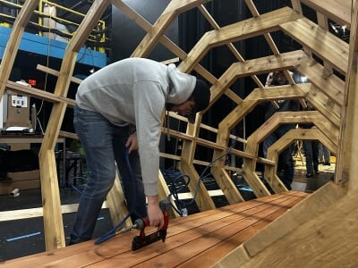 A student stands inside the partially constructed framework using a nail gun.