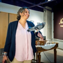 Smitha Rao poses with Sonnet, a pet Danzig Highflier pigeon on a leash. Behind, a red wall reads 'Nanowonder: Images of the Microscopic World.'