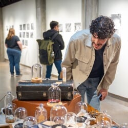 Gallery attendees closely observe small items under glass displays, including a toy car, butterfly wing, and scraps of fabric.