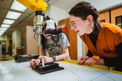 Two students lean over a lab table, examining a robotic arm.