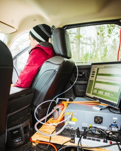 A computer, display, power source, and wires connecting it all are fashioned to a wood board in the backseat of a truck, with Ben Kopec in a red coat and dark hat seated looking forward in the front passenger seat.