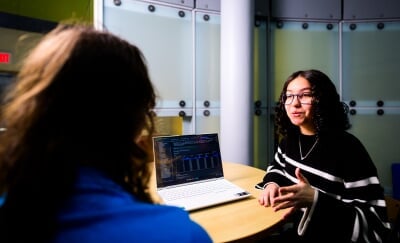 Diana Shadibaeva sits at a round table with a laptop displaying code on the table near her while she talks to another person.