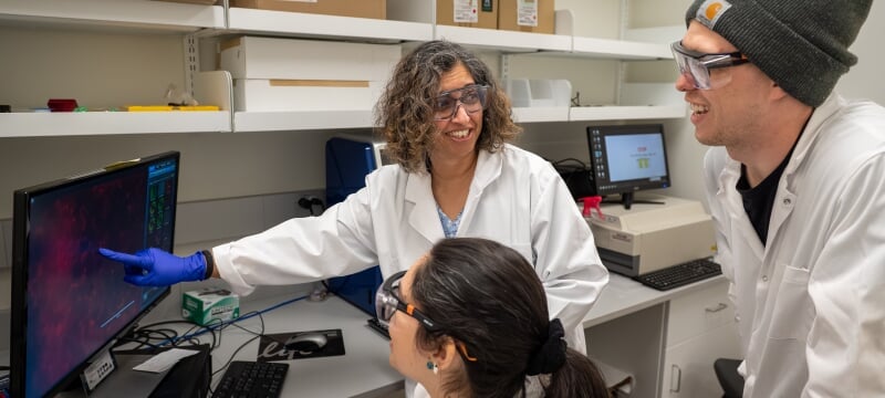 Smitha Rao and two of her research assistants in white lab coats analyze data on a computer screen in a laboratory setting.