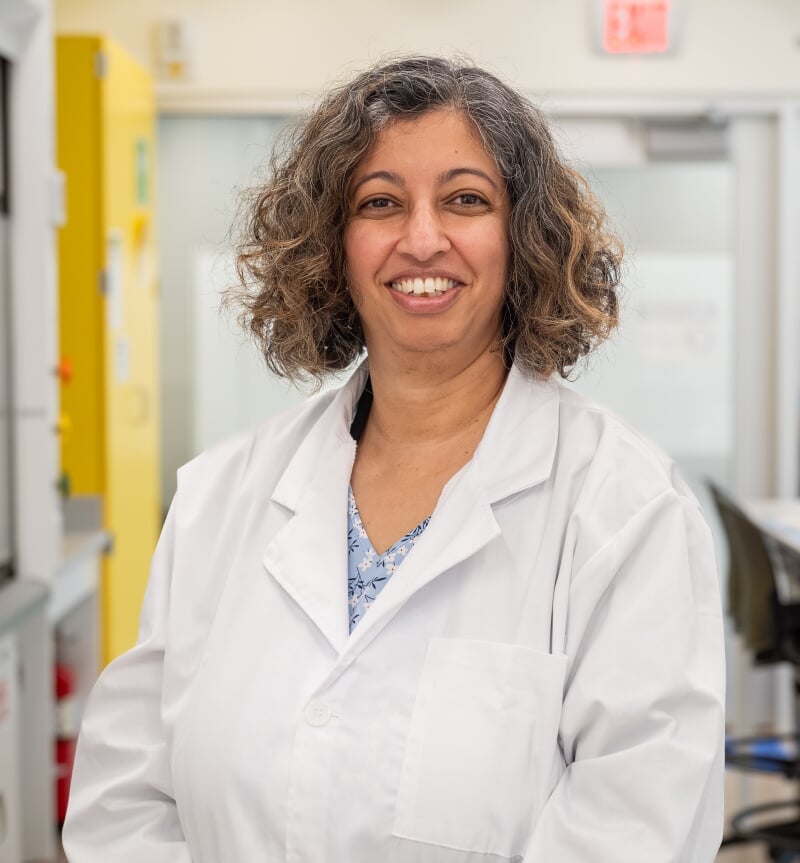 Smitha Rao wearing a white lab coat and blue gloves standing in a laboratory setting with equipment in the background.