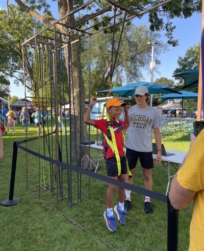 Child wearing a safety harness uses a steel hypogravity simulator at an outdoor event with an adult supervising nearby