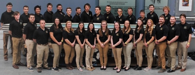 a team in black photo shirts and tan khakis stands around a long light-blue canoe on a stand in a gymnasium