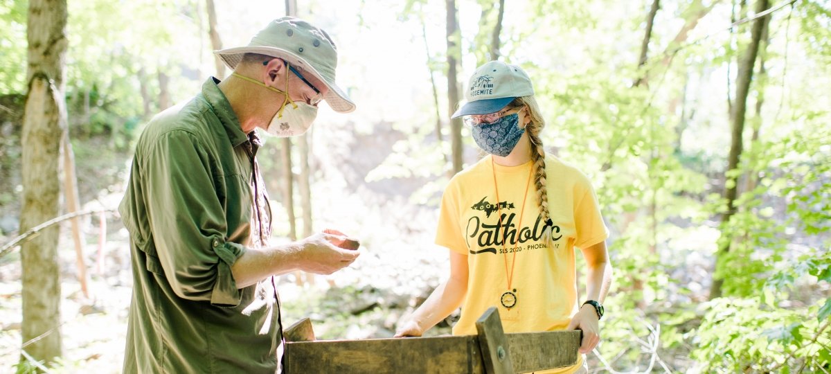 Two people wearing face coverings use a sluice box to sort through rocks and dirt.